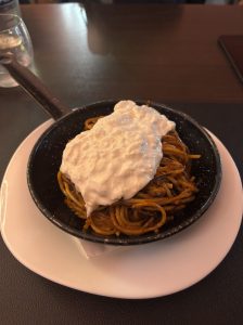 Spaghetti served in a small black skillet, topped with a generous portion of creamy burrata cheese